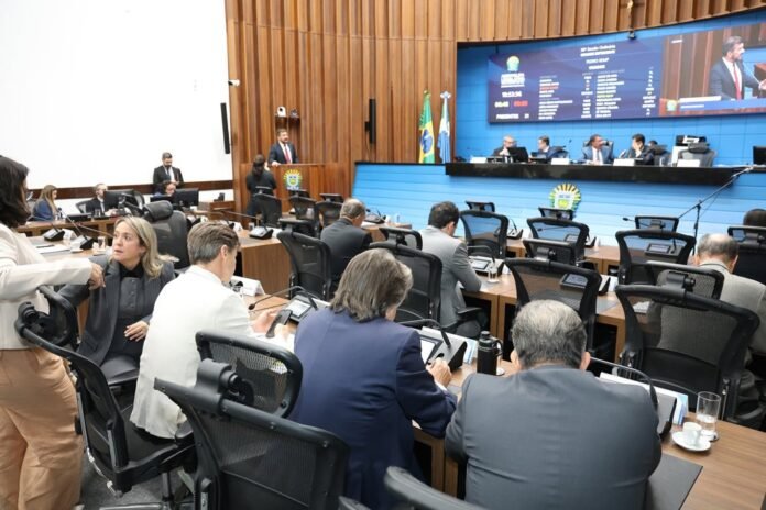 Formal government meeting in a wood-paneled chamber with attendees at desks and a blue display behind the panelists at the front of the room.