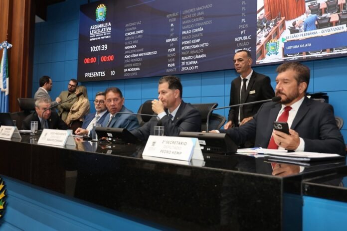 Panel of officials seated at a glossy black desk in a legislative chamber, with large screens displaying names and session info behind them