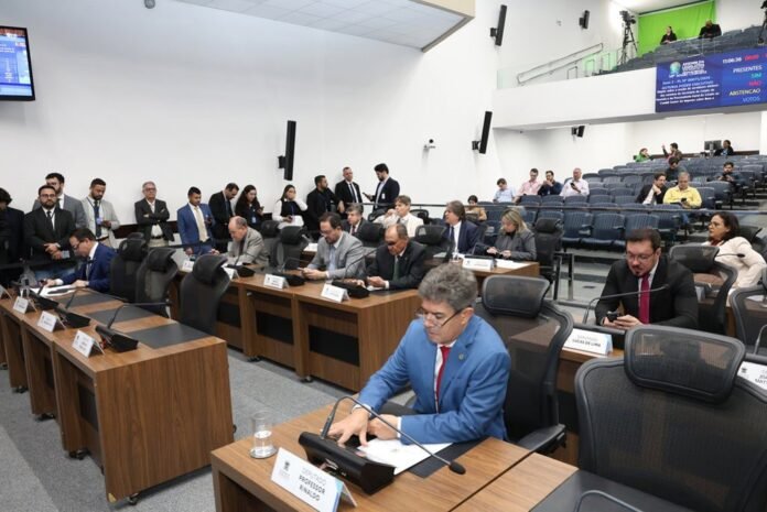 Parliamentary hearing with officials seated at desks, microphones and nameplates in a formal chamber