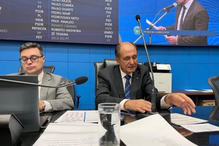 Two men in suits sit at a conference table with microphones, papers, and a glass of water during a formal meeting in a blue-walled room.
