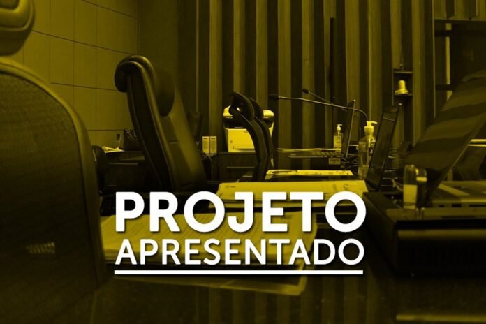 Office workspace with laptops and chairs at a long desk; bold white text on screen reads 'PROJETO APRESENTADO', indicating a project presentation.