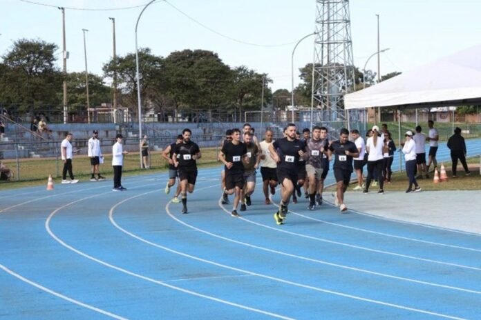Group of runners with bib numbers start a race on a blue outdoor track, with spectators and officials nearby.
