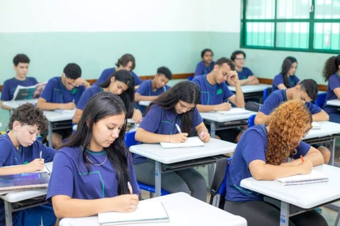 Students in matching purple shirts sit at desks in a bright classroom, writing in notebooks.