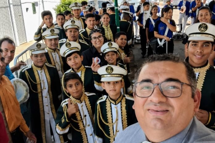 Selfie taken by a man with glasses in front, with a large group of marching band students in green and gold uniforms and hats behind him.