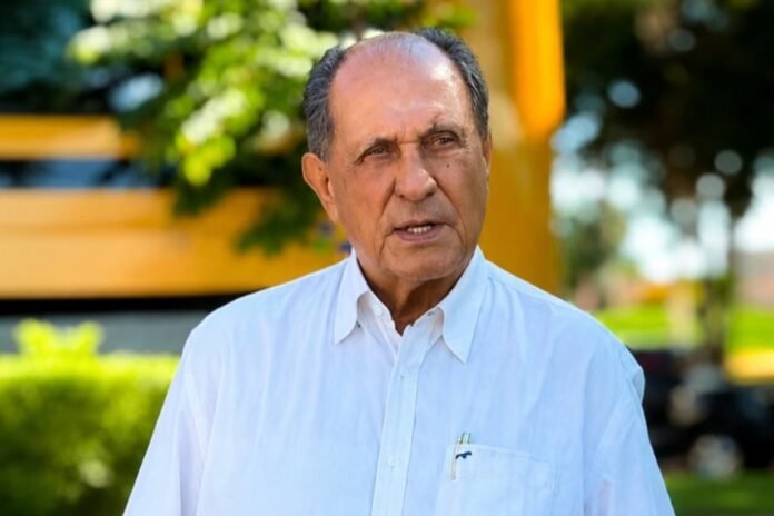 Older man in a white button-down shirt standing outdoors, talking with a blurred yellow structure and trees in the background.