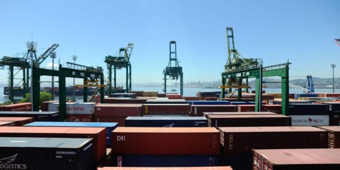Shipping port with stacked colorful containers, towering green gantry cranes, and a calm harbor beyond the water. City skyline visible in the distance.