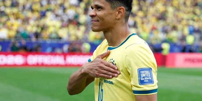 Brazilian footballer in a yellow Brazil jersey places his hand over his heart on the field, with a cheering crowd in yellow behind him.