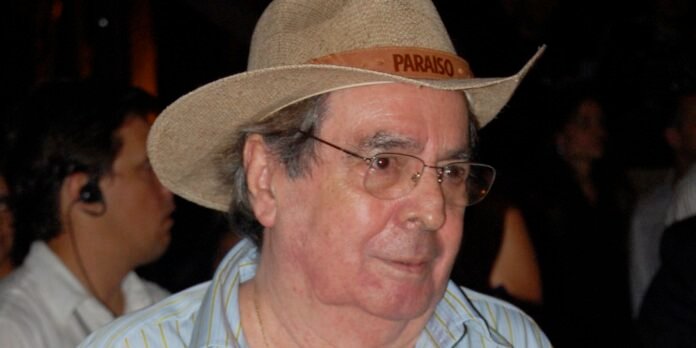 Older man in a straw hat with a 'PARAISO' band and glasses, looking to the side at a nighttime outdoor event.