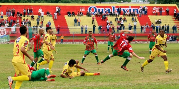 Soccer match with players in yellow and red-green uniforms chasing the ball on a grassy field, crowd in colorful stadium stands behind.