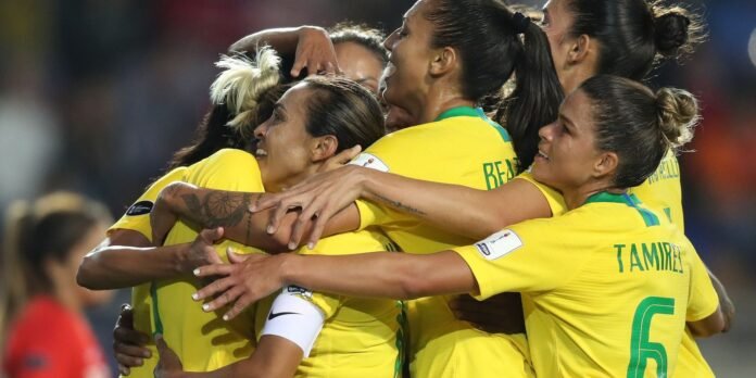 Brazilian women soccer players in yellow jerseys celebrate in a group hug on the field, toasting a goal.