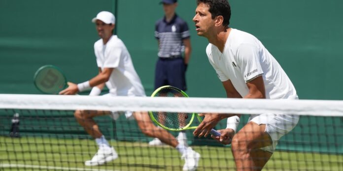Two male tennis players in white outfits chase the ball on a grass court, nets visible in foreground.