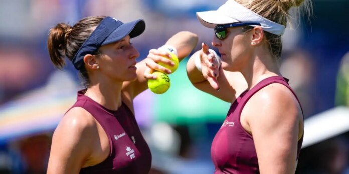 Two female tennis players in maroon tops and visors face off on a sunny court, one holding a tennis ball between them as they talk.