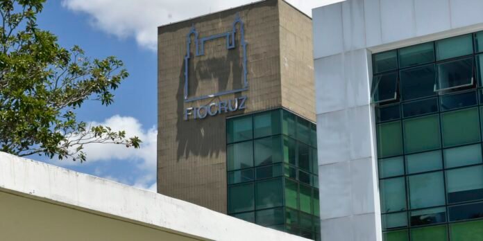 Modern building facade with green-tinted glass and a beige tower displaying the blue Fiocruz logo against a blue sky.