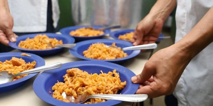 People scooping pasta into blue bowls in a cafeteria line, ready to serve meals.