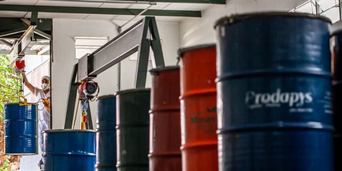 Worker in white PPE and mask uses a hoist to lift a blue drum in an industrial workshop with stacked barrels nearby
