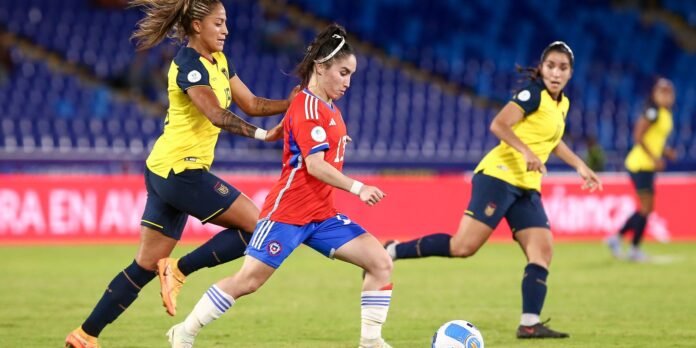 Female soccer player in a red jersey dribbles the ball while two opponents in yellow chase closely on a green field inside a stadium.