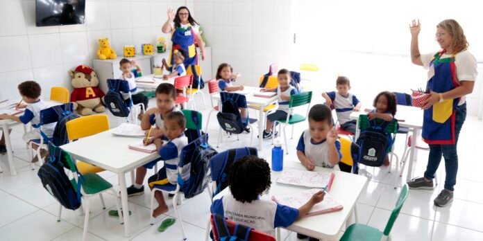 Bright classroom with about a dozen preschoolers in blue-and-white uniforms seated at white tables, drawing in notebooks. Backpacks hang on chairs, colorful chairs around the room, and a large stuffed bear and toy blocks sit on a cabinet. A teacher in a multicolored apron waves at the camera on the left; another teacher in a similar apron stands by the right, holding a container of supplies and raising a hand.