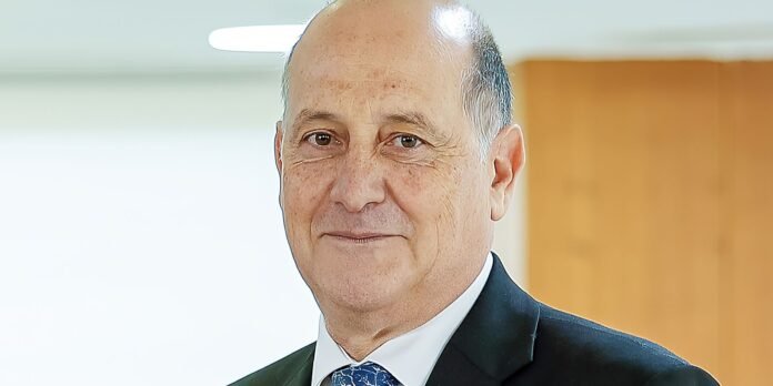Smiling older man in a dark suit and blue patterned tie, indoors in an office setting.
