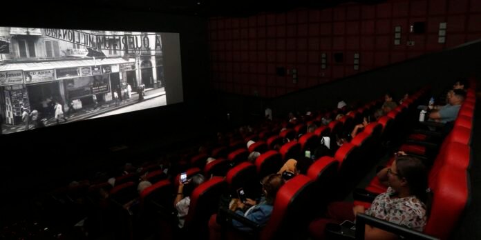 Audience seated in red upholstered cinema hall watching a large black-and-white film projected on screen at front.