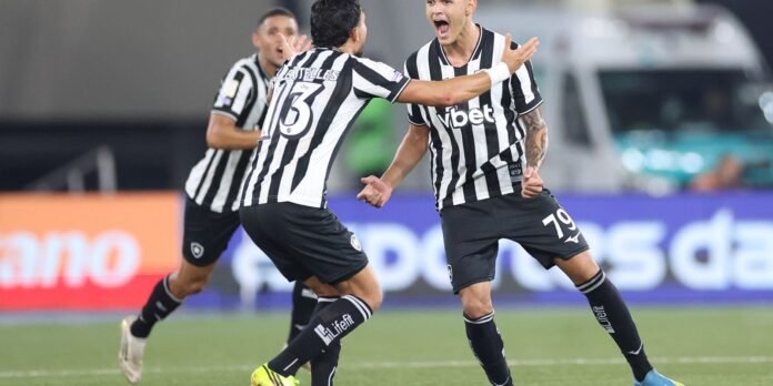 Two soccer players in black-and-white striped kits celebrate on the pitch, arms raised in triumph after a goal.