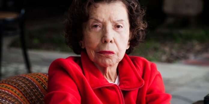 Elderly woman with short curly hair in a red jacket sits outdoors with a serious expression.