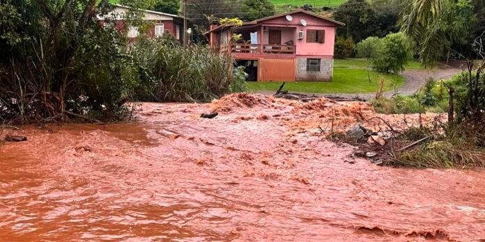 Reddish muddy floodwater fills a yard and riverbank near a pink two-story house with debris along the shore.