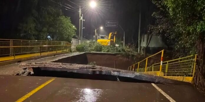 Nighttime street with a collapsed roadway blocking traffic; a trench spans the lane with broken asphalt and safety rails nearby, and a yellow excavator in the background while streetlights glow.