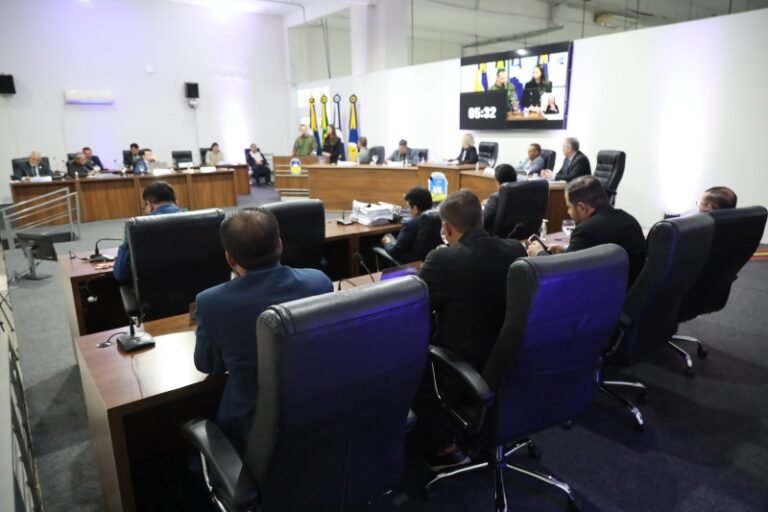 Formal government or city council meeting with officials around a U-shaped desk; a large screen shows video feeds and a timer, flags in the background.