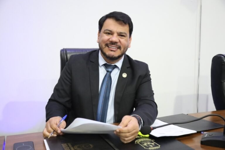Man in a dark suit and tie sits at a desk, smiling while reviewing documents in his hands.