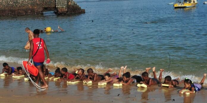 Lifeguard in a red shirt leads children practicing swimming with kickboards along the sunny beach shoreline.