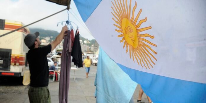 Man hangs clothes on a line under a canopy at a waterfront market, with a large Argentine flag featuring a sun in the foreground.