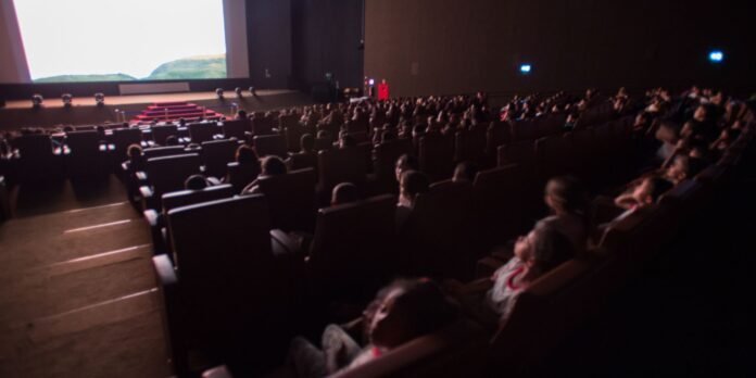 Audience seated in a dark auditorium watching a presentation on a large screen at the front of the room.