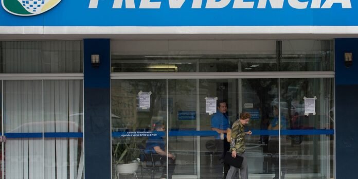 Front entrance of a bank with glass doors and blue signage; two people enter while a staff member in a blue shirt stands inside.