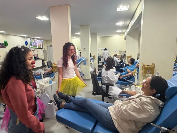 Two clowns entertain a donor in a bright clinic while she sits in a blue reclining chair to donate blood or receive care.