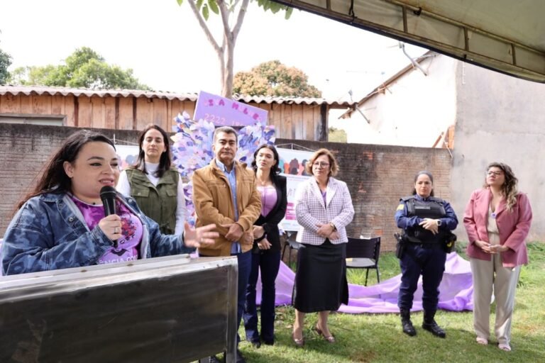 Woman speaking into a microphone at a podium during an outdoor community event, with six people standing behind her near a purple backdrop and brick wall.