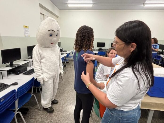 A school computer lab where a staff member administers a shot to a student as a large white mascot watches nearby.