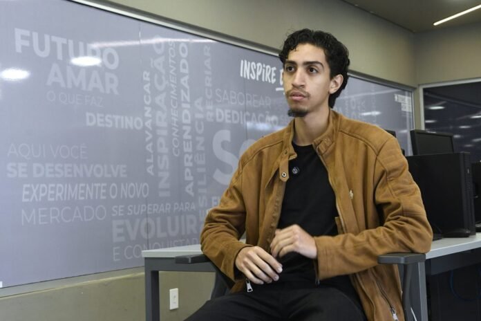 Young man in a brown jacket sits at a desk in a modern office, looking thoughtful.