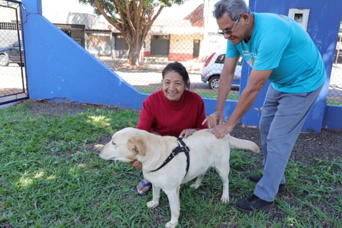 Smiling woman in red kneels with a light-colored dog while a man in blue pets the dog outdoors.