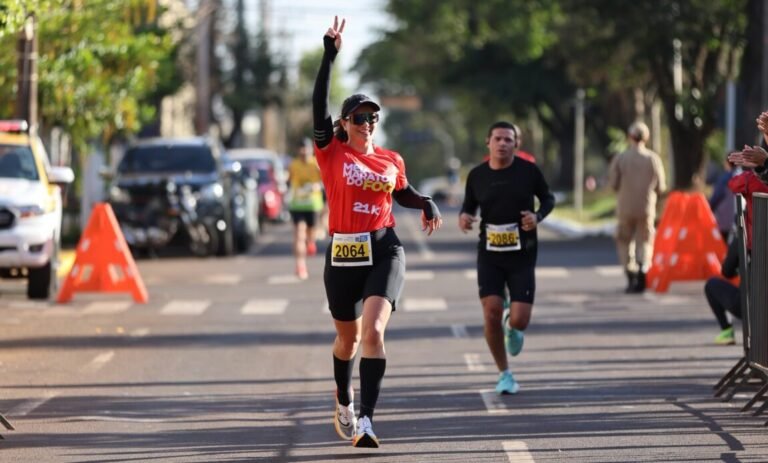 Female runner in a red shirt raises two fingers in a peace sign as she crosses the finish line, bib 2064.