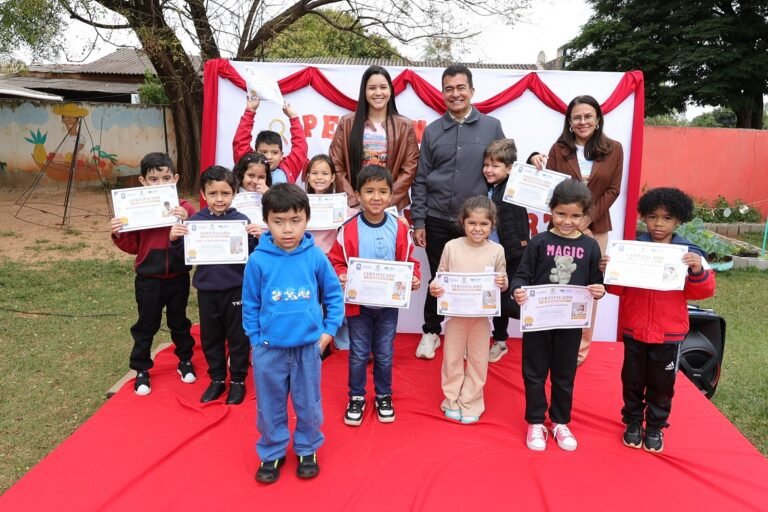 Group of children posing on a red carpet with certificates, flanked by adults outdoors during an award ceremony.