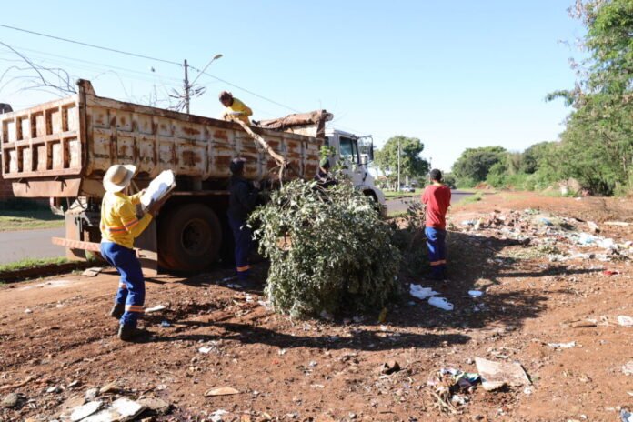 Workers load a large tree branch into a rusty dump truck during roadside cleanup on a sunny day fronting a dirt lot and littered ground.