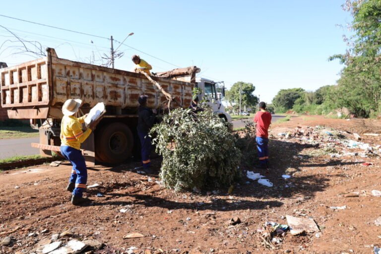Workers load a large tree branch into a rusty dump truck during roadside cleanup on a sunny day fronting a dirt lot and littered ground.
