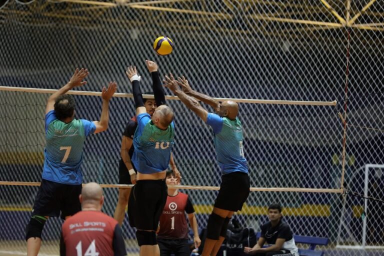 Indoor volleyball game: four players jump at the net to block a ball, blue jerseys and knee pads, with a yellow volleyball overhead.