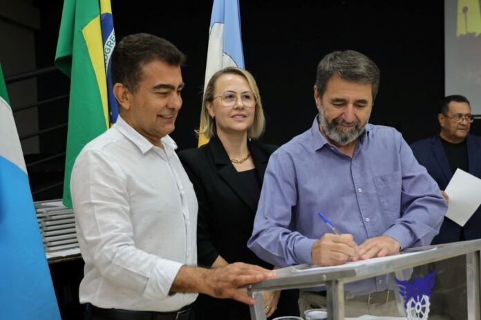 Three adults at a signing event: a man in a white shirt and a man in a light blue shirt signing documents at a podium, with a woman in a black blazer watching, flags in the background.