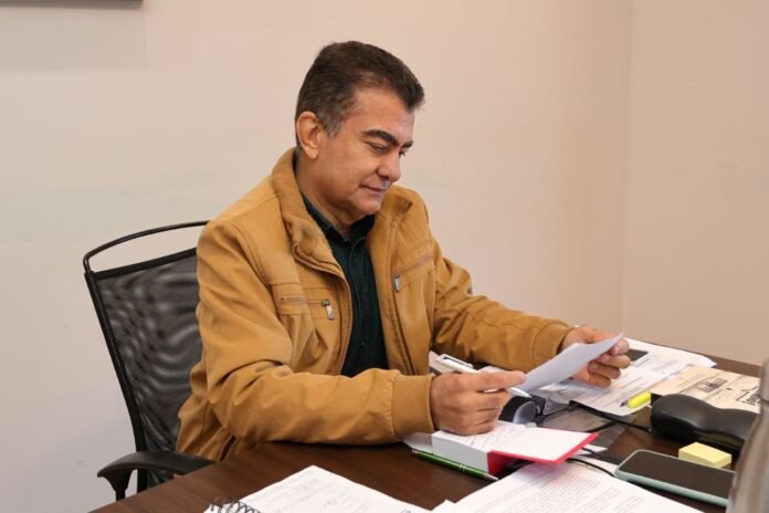 Man in a tan jacket sits at a desk, signing or reviewing papers surrounded by office supplies.