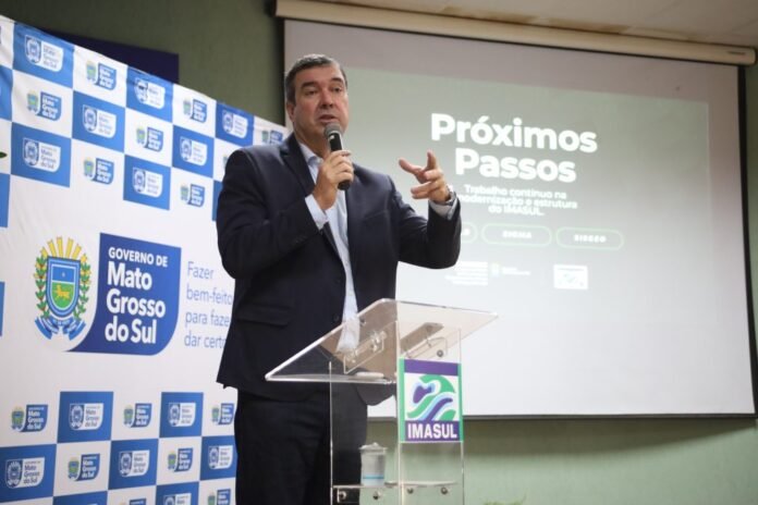 Man in a dark suit speaks into a microphone at a clear podium during a government event, projector displays 'Próximos Passos'