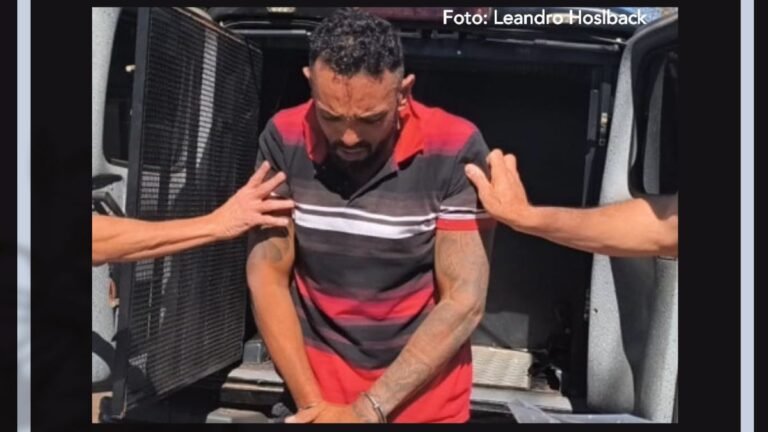 Man in a red and black striped polo is restrained by two people at the back of a van.