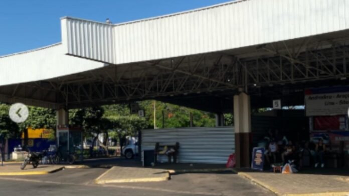 Open-air shelter with a large white metal roof; people sit along a shaded area at a market or station, with a motorcycle parked nearby and trees in the background.