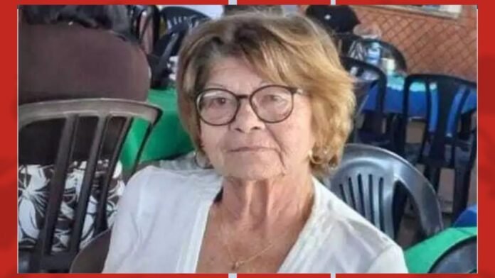 Portrait of an older woman with short blonde hair, round glasses, and a white blouse, seated at an outdoor event.