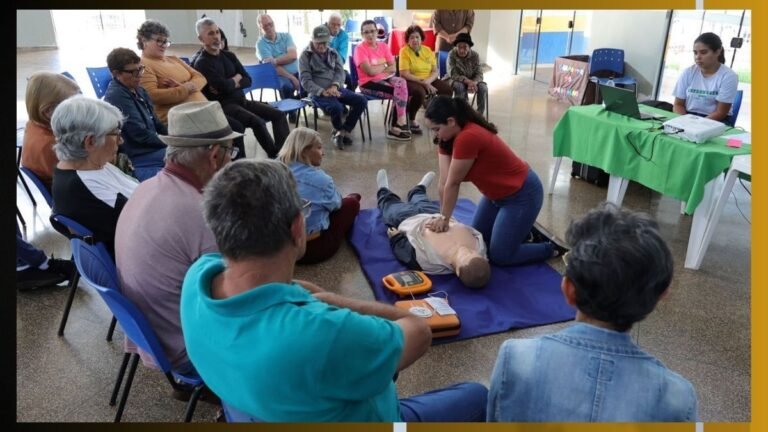 CPR training session: a woman performs chest compressions on a manikin while a group watches in a circle in a bright room. An AED device sits nearby, instructor at front.